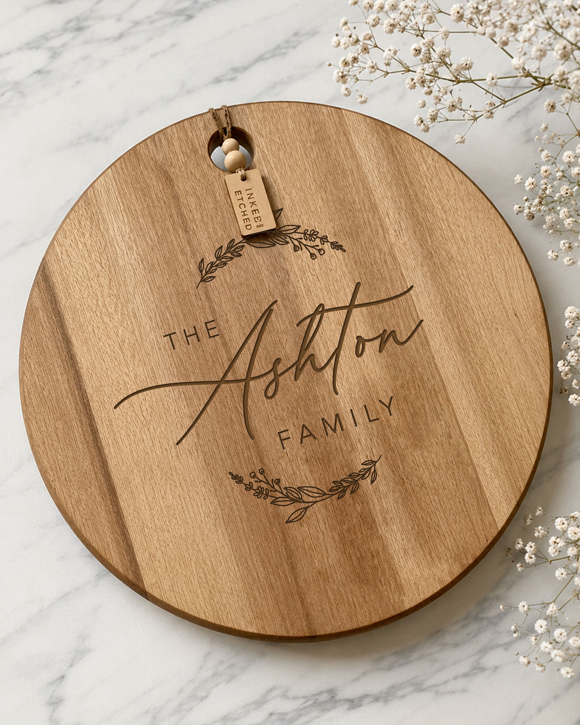 Personalised engraved acacia cheeseboard with custom family name, photographed on a marble surface with baby's breath flowers.