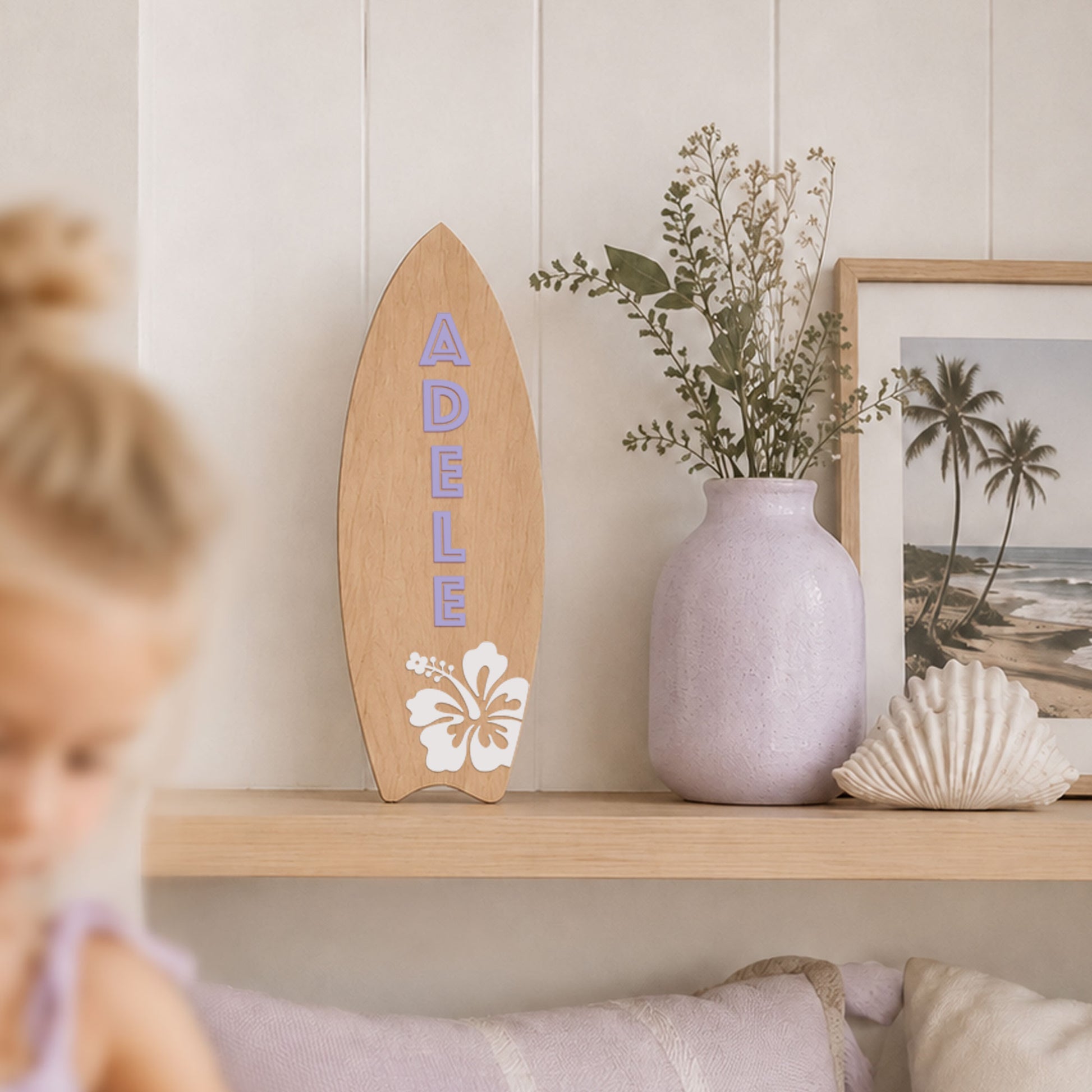 Personalised wooden surfboard wall decor with lavender name Adele and white hibiscus flower displayed on a shelf in a coastal girls bedroom.