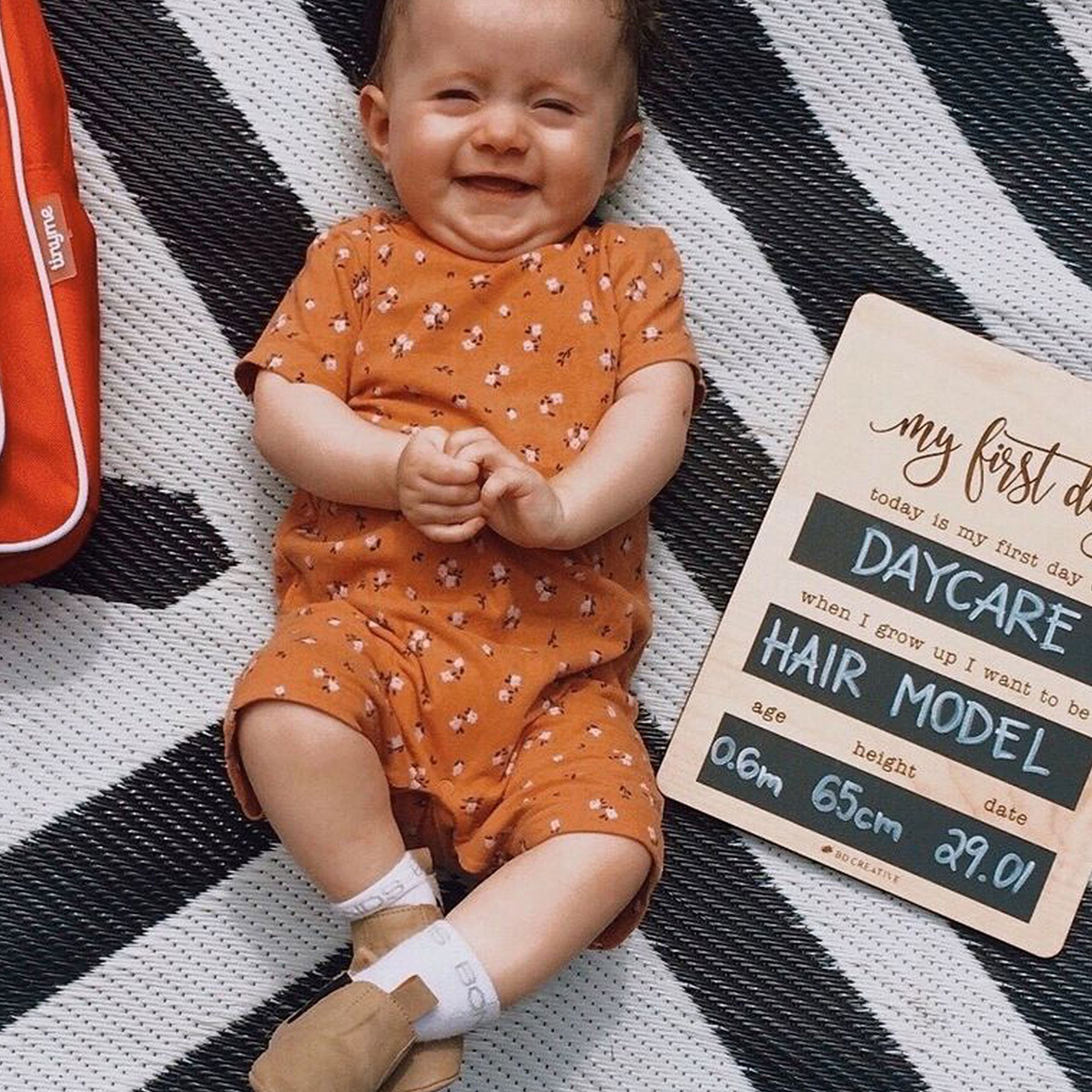 Smiling baby lying beside a reusable wooden first day of daycare milestone board.