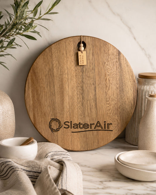 Branded round acacia serving board engraved with the SlaterAir company logo displayed in a modern kitchen.