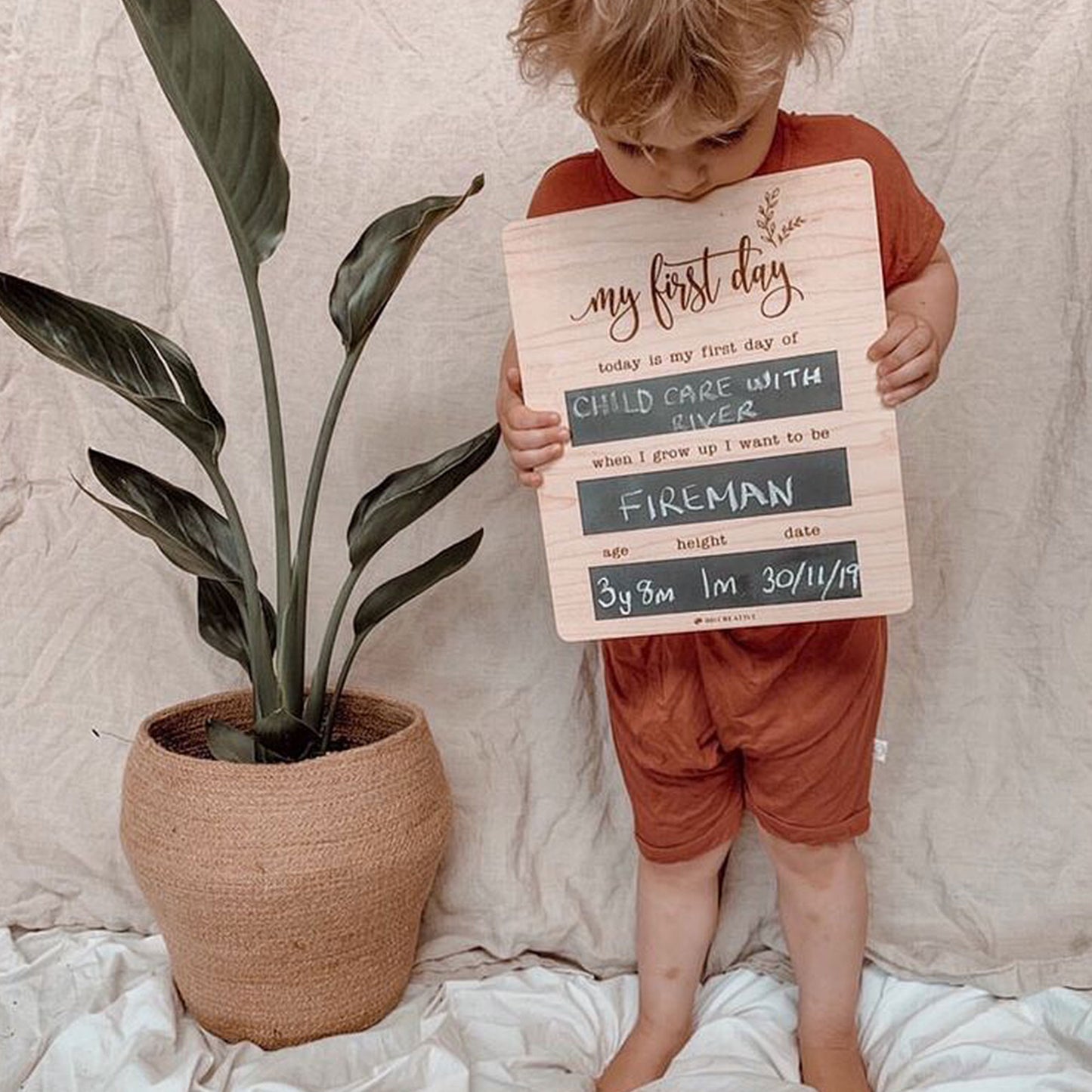 Young boy holding a reusable wooden first day of childcare chalkboard beside a potted plant.
