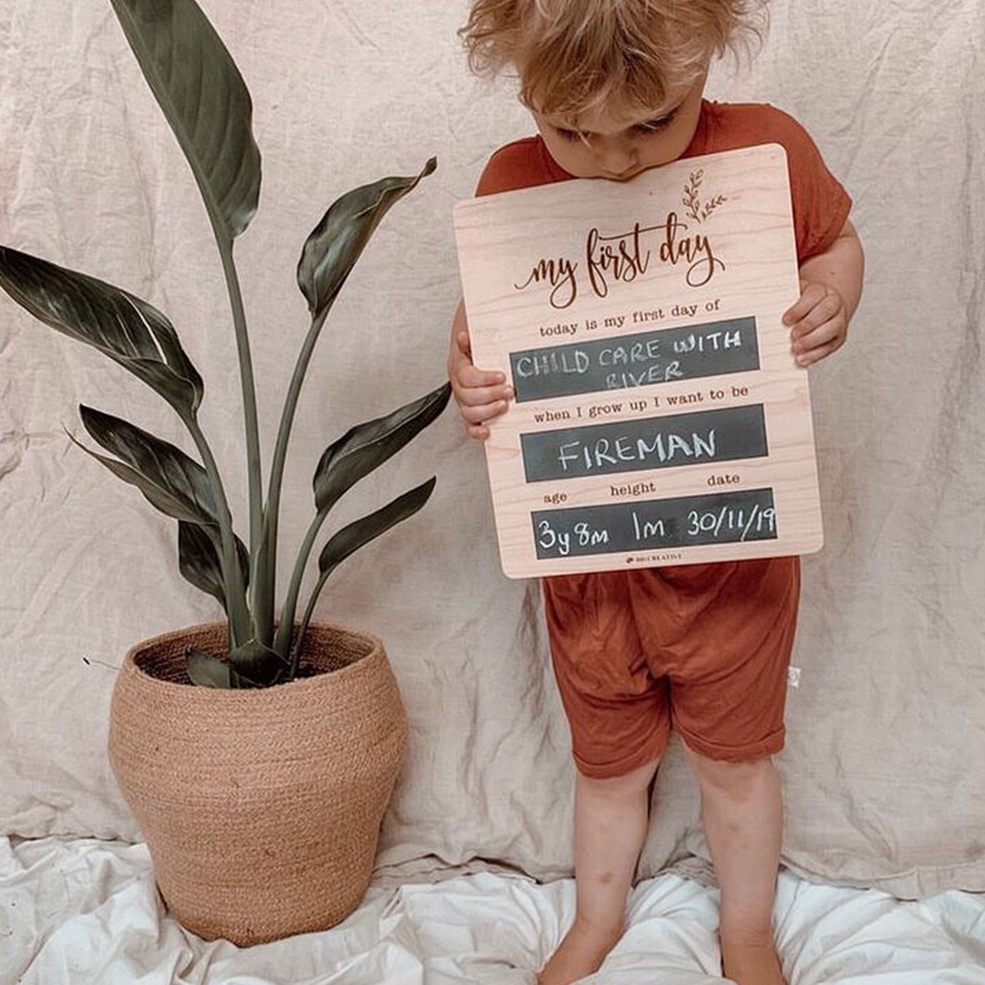 Young boy holding a reusable wooden first day of childcare chalkboard beside a potted plant.