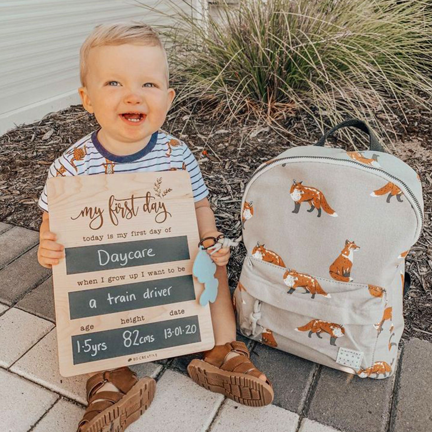 Smiling toddler beside a backpack holding a reusable wooden first day of daycare chalkboard sign.
