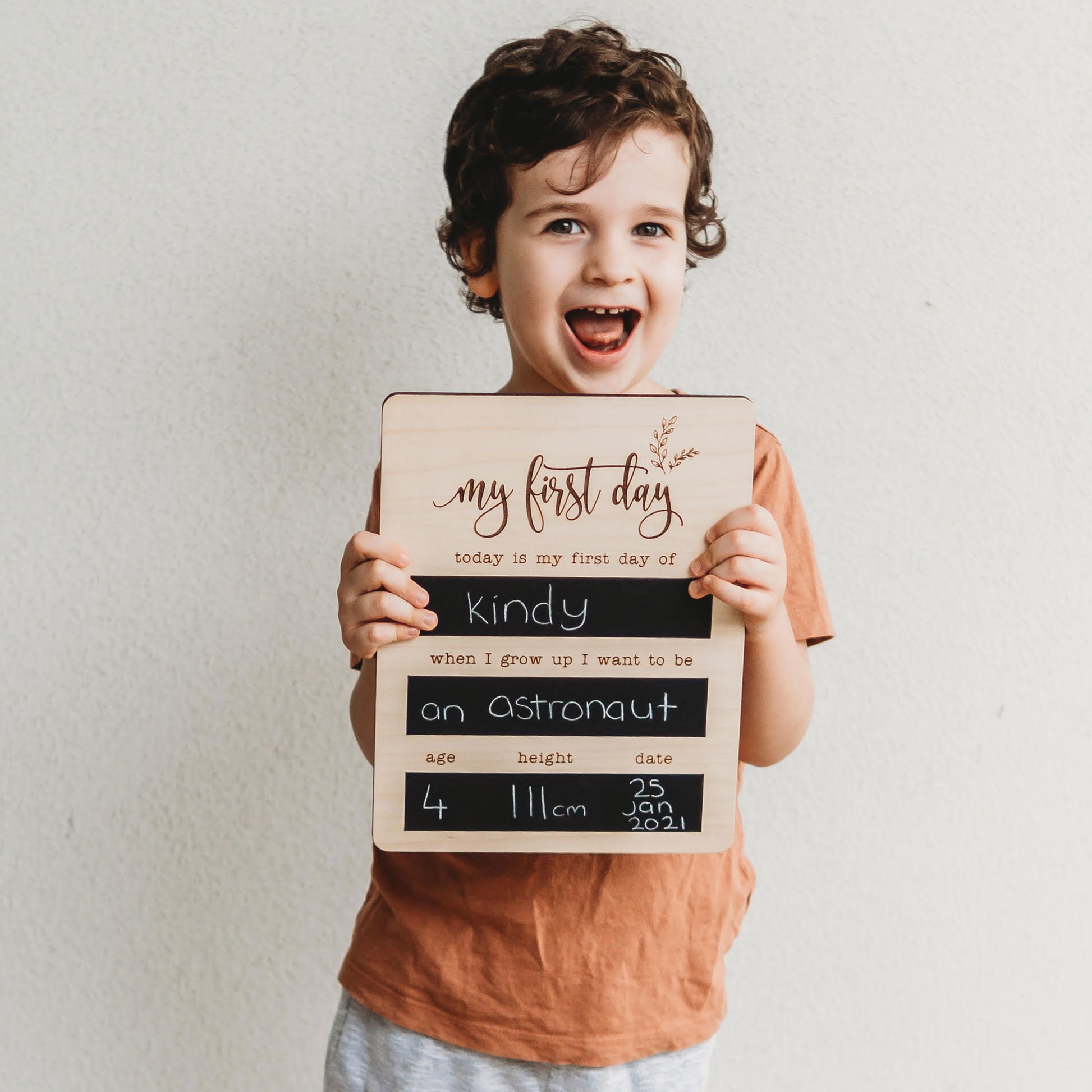 Smiling boy holding a reusable wooden first day of kindy milestone board with astronaut ambition.