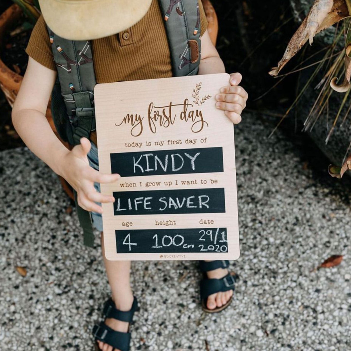 Child wearing a backpack and holding a reusable wooden first day of kindy chalkboard.