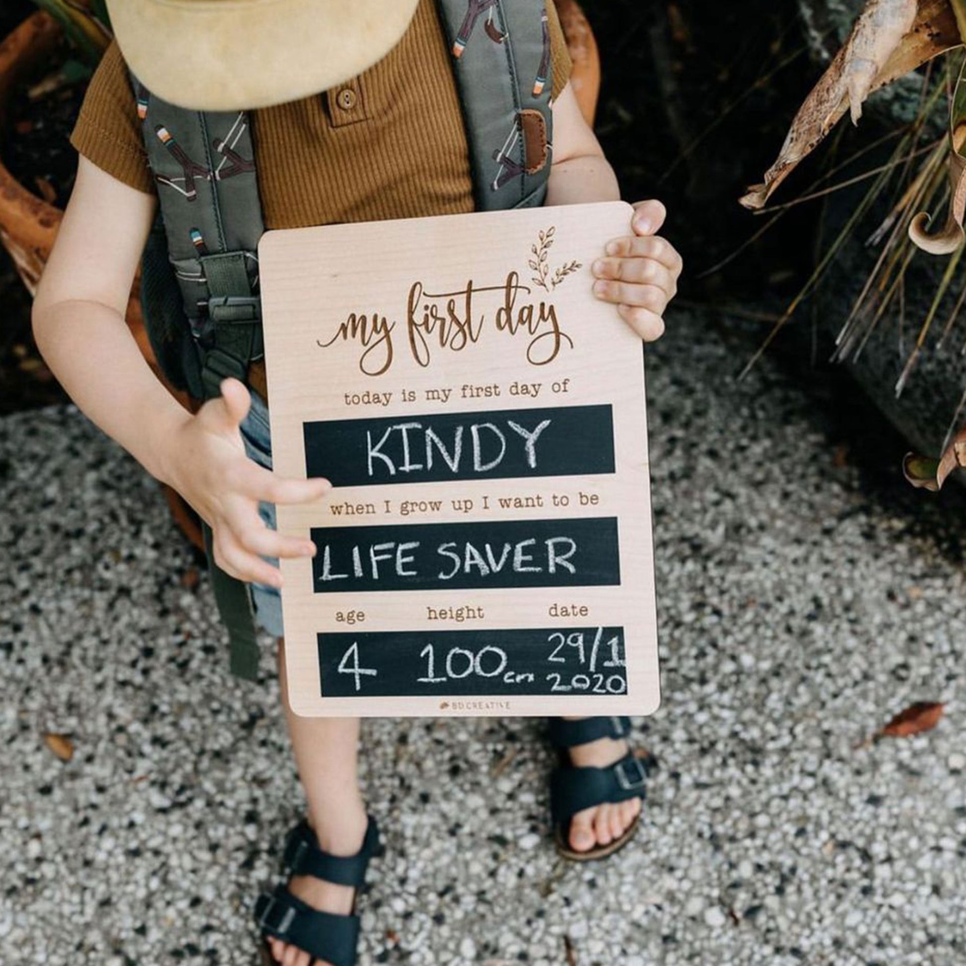 Child wearing a backpack and holding a reusable wooden first day of kindy chalkboard.
