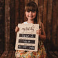 Girl holding a reusable wooden first day of school chalkboard filled in for Year 2.
