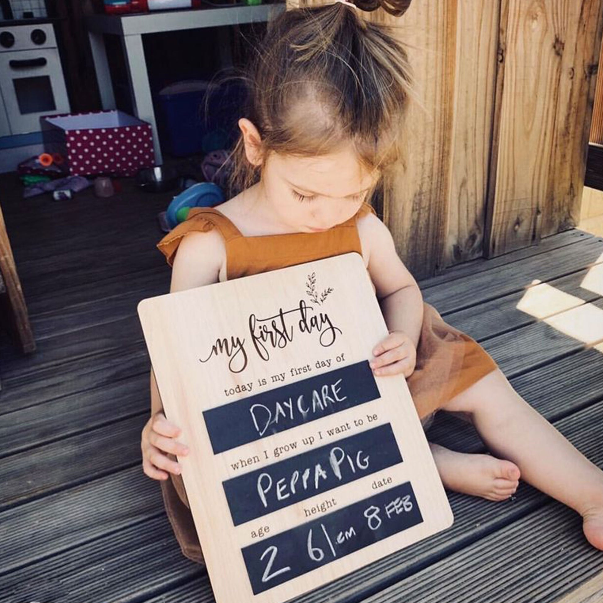 Young girl seated outdoors holding a reusable wooden first day of daycare chalkboard sign.