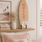 Large personalised wooden surfboard wall decor with hibiscus flower displayed on a shelf in a coastal style children's bedroom. The surfboard is on a shelf above a bed.