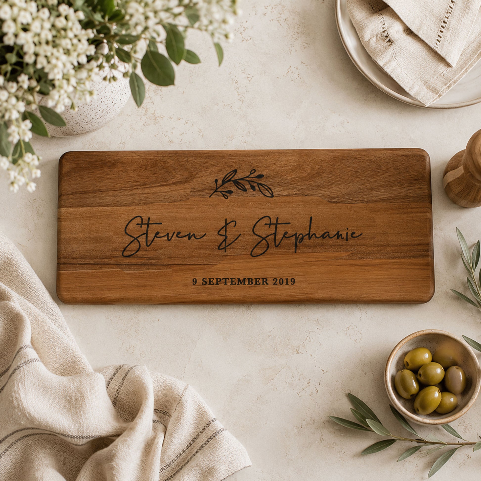 ersonalised wooden serving board engraved with names and wedding date displayed in a neutral flat lay with olives and linen.