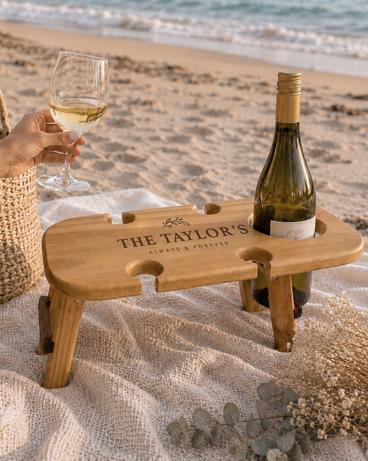 Personalised foldable wooden wine picnic table on the beach with a wine bottle, wine glass and picnic blanket, perfect for outdoor wine and cheese picnics.