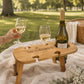 Personalised engraved wooden wine picnic table in a park holding a wine bottle and two wine glasses on a picnic blanket beside a picnic basket.