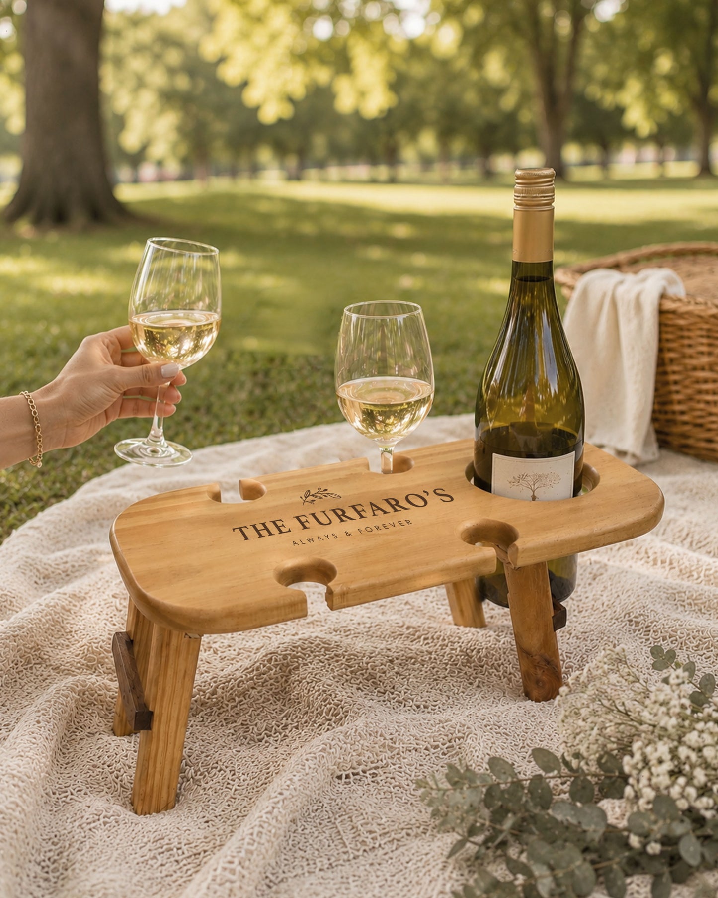 Personalised engraved wooden wine picnic table in a park holding a wine bottle and two wine glasses on a picnic blanket beside a picnic basket.