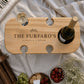 Personalised wooden wine picnic table engraved with a family name, holding a wine bottle and wine glass, styled with cheese, crackers, grapes and strawberries on a picnic blanket.