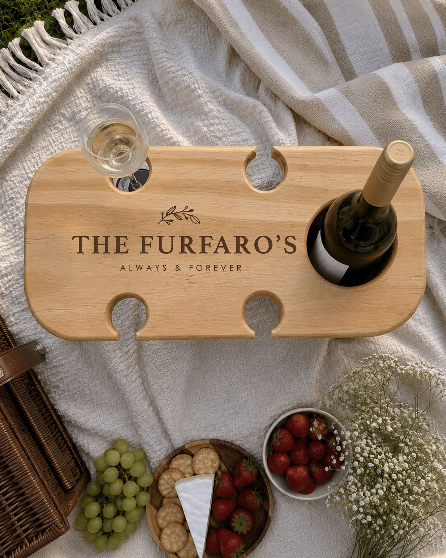 Personalised wooden wine picnic table engraved with a family name, holding a wine bottle and wine glass, styled with cheese, crackers, grapes and strawberries on a picnic blanket.