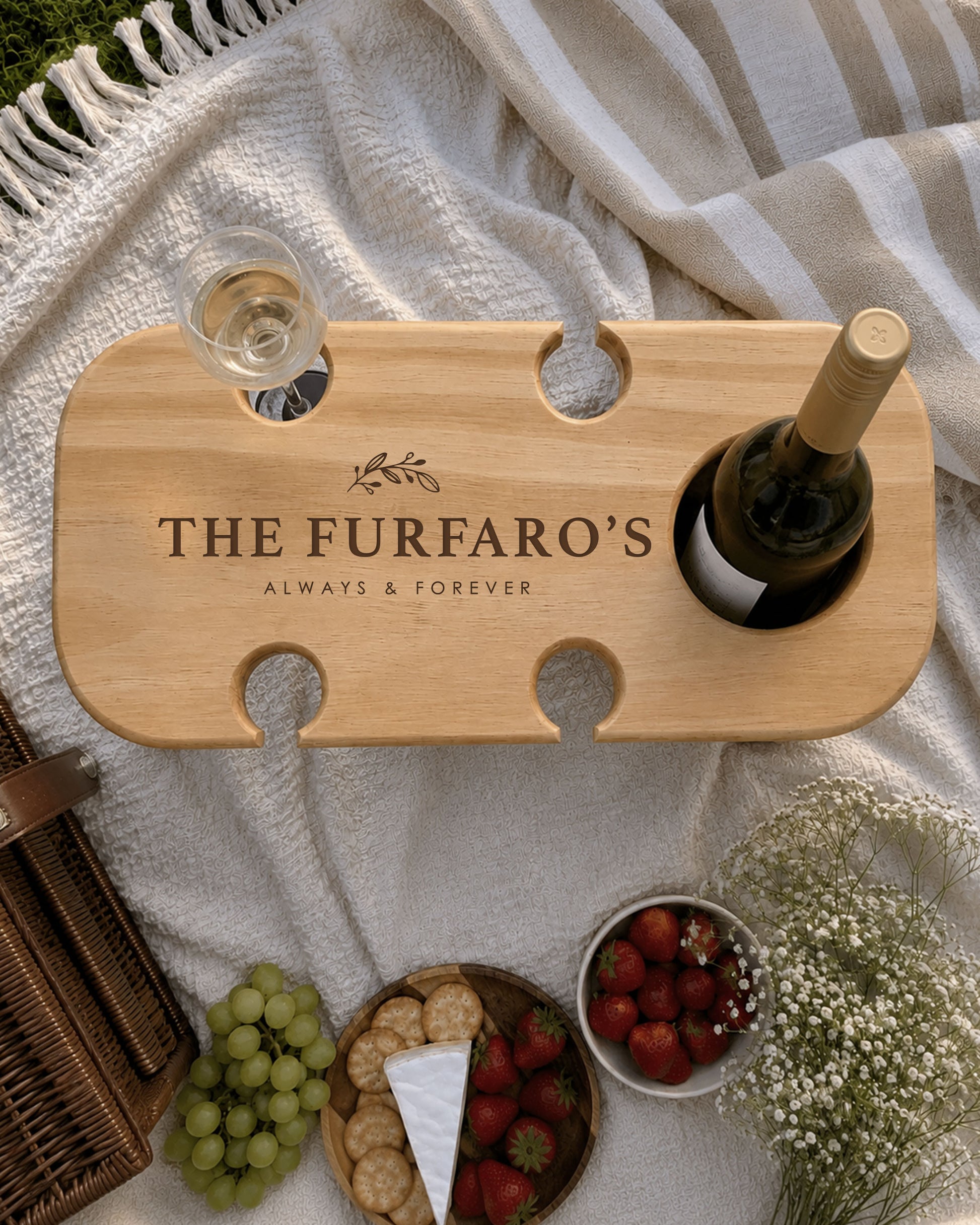 Personalised wooden wine picnic table engraved with a family name, holding a wine bottle and wine glass, styled with cheese, crackers, grapes and strawberries on a picnic blanket.