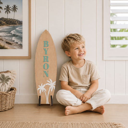 Personalised wooden surfboard wall decor with the name Byron, featuring white palm trees and a natural timber finish, styled in a modern coastal boys bedroom with beach-inspired décor.