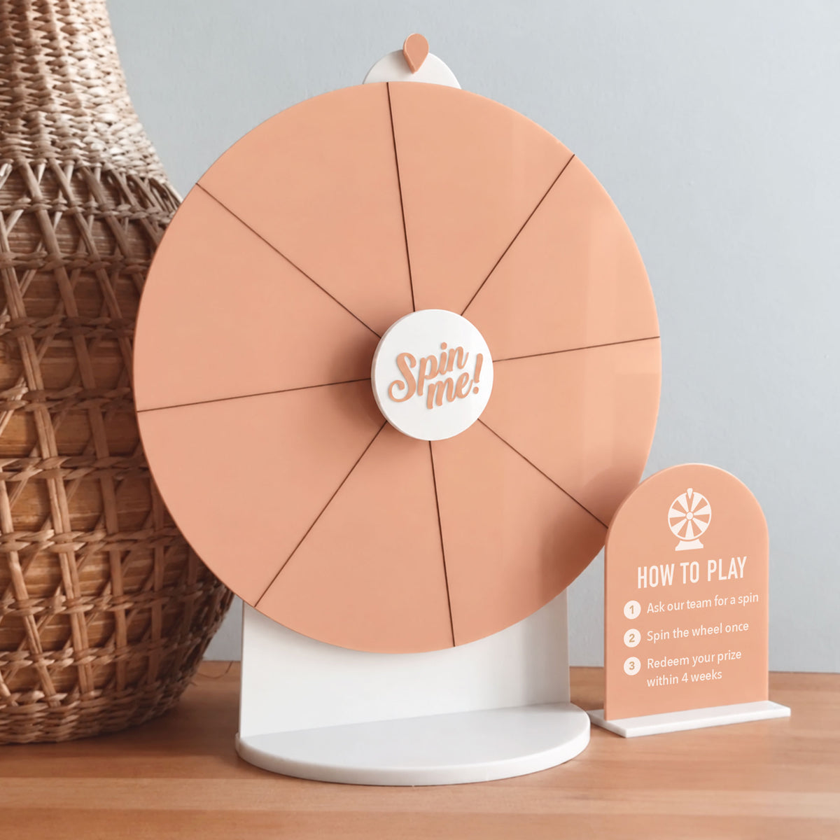 Angled view of a peach acrylic Spin The Wheel with a white stand, “Spin Me” centre and matching instructions sign.
