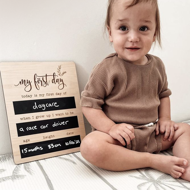 Smiling toddler sitting beside a reusable wooden first day of daycare milestone board.